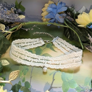 Pu&ograve; includere: Girocollo in pizzo color avorio con un delicato motivo floreale. La collana presenta una catena e una chiusura argentate e una piccola perla finta pendente. Lo sfondo comprende vegetazione e fiori blu e gialli.