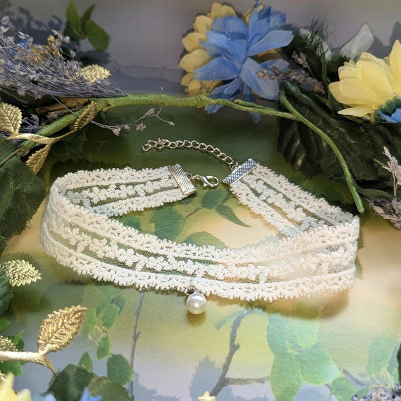 Pu&ograve; includere: Girocollo in pizzo color avorio con un delicato motivo floreale. La collana presenta una catena e una chiusura argentate e una piccola perla finta pendente. Lo sfondo comprende vegetazione e fiori blu e gialli.