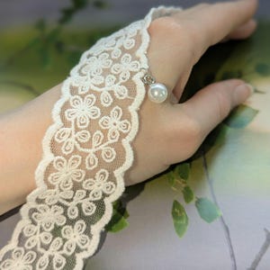 Pu&ograve; includere: Bracciale da dito in pizzo avorio con motivo floreale e bordi smerlati. Una piccola perla bianca pende dal bracciale tramite un anello color argento. Il bracciale &egrave; progettato per essere indossato sulla mano.