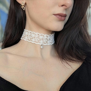 Pu&ograve; includere: Una collana girocollo in pizzo bianco con un motivo floreale e un piccolo pendente a perla. La collana &egrave; indossata intorno al collo. Sono visibili anche orecchini dorati a forma di foglia.