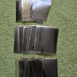 May include: A collection of rectangular, semi-transparent dark gray film negatives arranged in three distinct horizontal stacks. Each individual piece of film is thin, flexible, and features a smooth, glossy surface with varying levels of opacity.