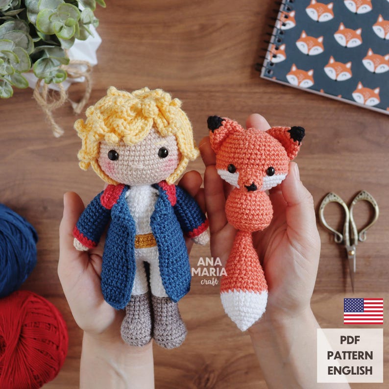 May include: Crocheted doll of a boy in a blue jacket with red trim and a crocheted fox. The boy doll has blonde hair and is wearing a white shirt. The fox is orange with a white belly and tail tip. The image includes the text "ANA MARIA craft" and "PDF PATTERN ENGLISH".