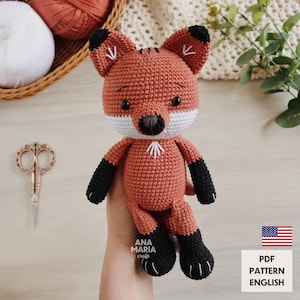 Puede incluir: Un juguete de zorro tejido a ganchillo en tonos naranja, blanco y negro. El zorro tiene un pecho blanco, patas negras y una nariz negra. La imagen también incluye el texto "ANA MARIA craft" y "PDF PATTERN ENGLISH".