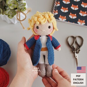 May include: A handmade crochet doll of a character with yellow hair, wearing a blue jacket with red trim, white pants, and gray boots. The doll is held in hands. The doll is a PDF pattern from Ana Maria Craft.