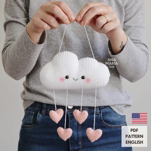 Könnte beinhalten: Eine weiße Häkelwolken-Mobile mit einem lächelnden Gesicht und rosa Wangen, mit fünf rosa gehäkelten Herzen, die darunter hängen. Die Mobile wird von weißen Schnüren gehalten. Der Text "ANA MARIA craft" ist sichtbar.
