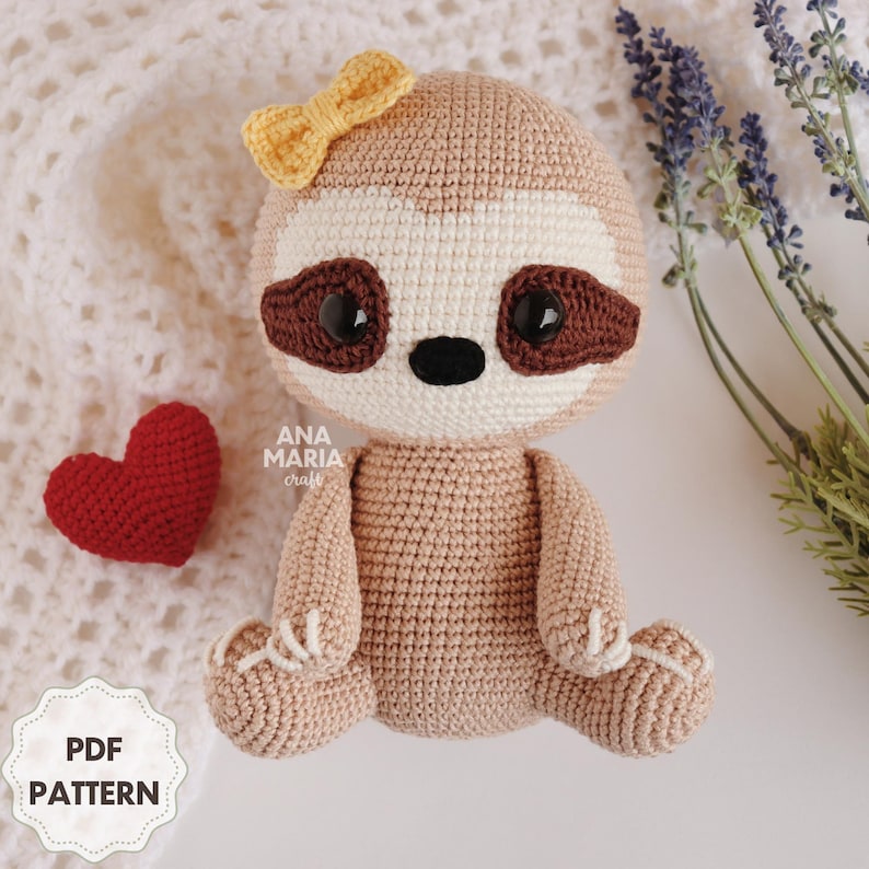May include: A crocheted sloth amigurumi toy with brown eyes, a white nose, and a yellow bow. The sloth is sitting with its arms and legs outstretched.