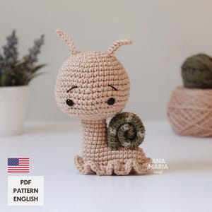 Puede incluir: Una figura de caracol de ganchillo en beige con una concha verde. El caracol tiene ojos negros, mejillas rosadas y una expresión sonriente. La base del caracol tiene un borde con volantes. La imagen también incluye el texto "PDF PATTERN ENGLISH" y la bandera estadounidense.