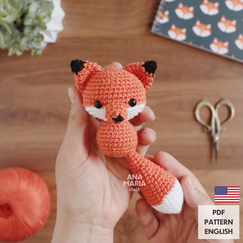 May include: A crocheted fox toy in shades of orange, white, and black. The fox has a rounded body, a long tail with a white tip, and black-tipped ears. The toy is handmade and features the text "ANA MARIA craft".
