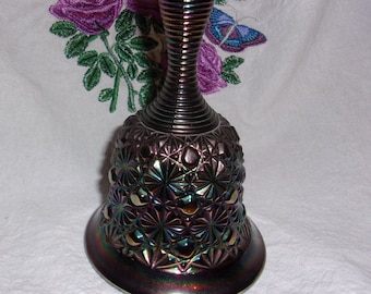 May include: A dark iridescent glass bell with a ribbed handle. The bell has a starburst pattern and is sitting on a white surface.