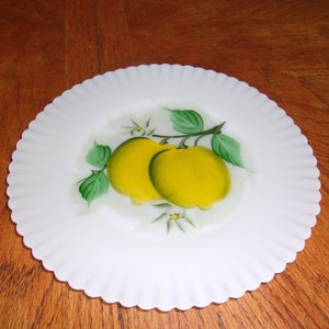 May include: White ceramic plate with a scalloped edge, featuring a hand-painted design of two yellow lemons with green leaves and white flowers.