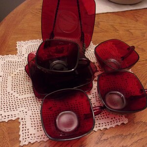 May include: A set of ruby red glass teacups and saucers. The square cups have handles and sit on matching square saucers. The set is arranged on a white lace doily on a wooden table. The cups and saucers are made of a textured glass.