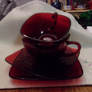 May include: A stack of three ruby red glass teacups and saucers. The cups have a square shape with a handle, and the saucers are also square. The set is placed on a white cloth with embroidered floral details.