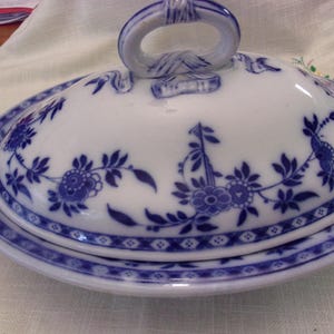 May include: Oval, lidded serving dish with a white base and blue floral design. The lid has a handle and is decorated with blue floral patterns. The rim of the dish has a blue geometric border. A classic piece for serving food.