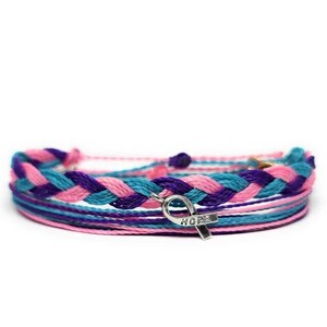 Peut inclure: Un bracelet multi-brins avec des cordons tressés roses, violets et bleus. Un charme argenté en forme de ruban avec le mot "HOPE" gravé dessus pend au bracelet.