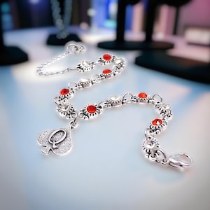 May include: Silver-toned bracelet with red and clear gemstones. The bracelet features a spade charm with the letter "Q". The gemstones are set in a floral design. The bracelet is likely made of a metal alloy.