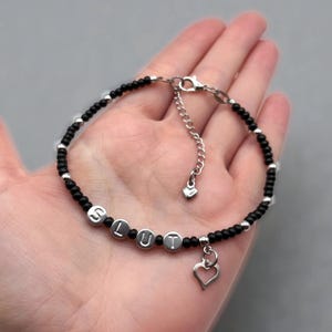 May include: A beaded bracelet featuring small black beads interspersed with silver-toned beads. Four silver-toned circular beads spell out the word SLUT. The bracelet includes a silver-toned heart charm and an adjustable chain with a small heart tag.
