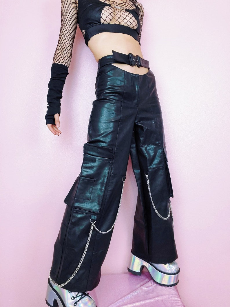 May include: Black leather pants with multiple cargo pockets and silver chain accents. The pants have a cut-out detail at the waist and are worn with a black mesh top and silver platform shoes.