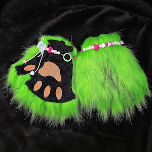May include: A pair of bright green furry fingerless gloves with black paw pads and brown paw prints. The gloves are decorated with beaded accents in pink, white, and green. The background is black.
