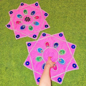 May include: Two pink, star-shaped decorative mats with a floral design. Each mat features a central flower and smaller flowers around the edges. The mats have a silver trim and are placed on a green surface.