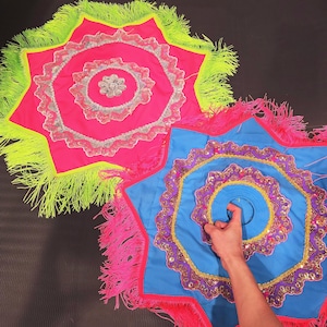 May include: Two star-shaped, decorative fabric pieces. One is hot pink with neon green fringe, the other is blue with pink fringe. Both have intricate silver and gold embellishments in concentric circles.