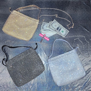 May include: Three sparkly, beaded handbags in gold, black, and silver, each with a different strap. A stack of dollar bills and a pink lipstick are also visible. The bags are square-shaped and appear to be evening bags.