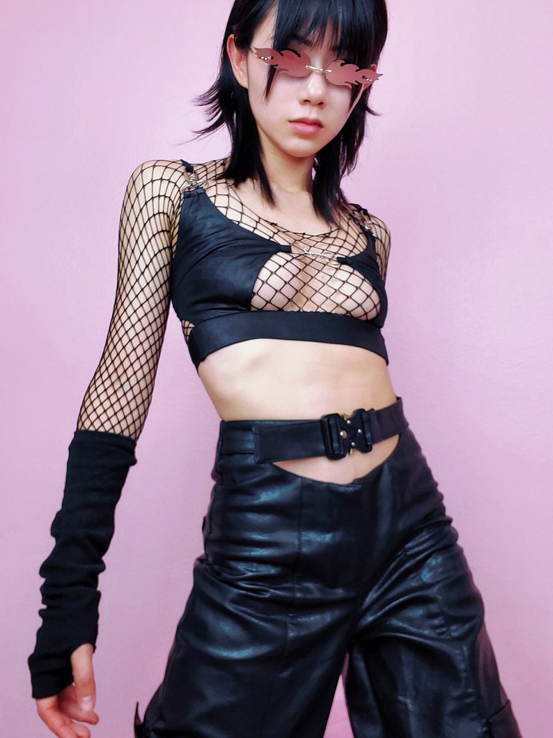May include: A person wearing a black mesh top with a cut-out design, black leather pants with a belt, and black fingerless gloves. The person is wearing pink sunglasses with a flame design.