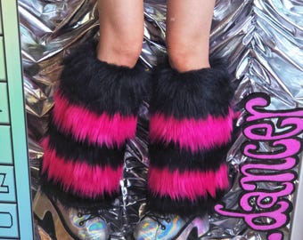 May include: A pair of leg warmers featuring horizontal stripes of black and hot pink faux fur. The leg warmers are worn over silver holographic platform boots with black laces, showcasing a thick, reflective sole and a textured metallic finish.