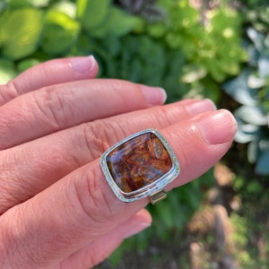 May include: A silver ring featuring a large, rectangular, cushion-cut gemstone with a polished surface. The stone displays a swirling pattern of deep brown, amber, and black tones. The metal setting has a textured, hammered edge finish.