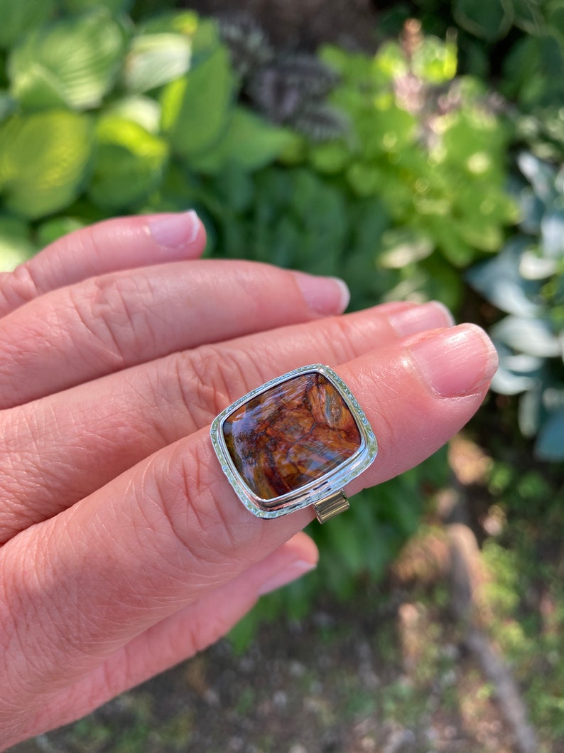 May include: A silver ring featuring a large, rectangular, cushion-cut gemstone with a polished surface. The stone displays a swirling pattern of deep brown, amber, and black tones. The metal setting has a textured, hammered edge finish.