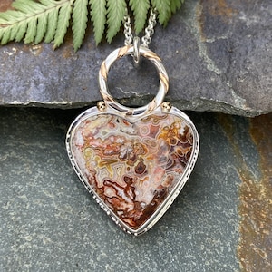 May include: A silver and gold heart-shaped pendant featuring a brown and white agate stone. The pendant is on a silver chain.