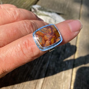 May include: A silver ring featuring a square-shaped, polished gemstone with a marbled pattern of deep brown, amber, and golden-orange hues. The metal setting has a textured, hammered edge surrounding the stone, creating a distinct border.