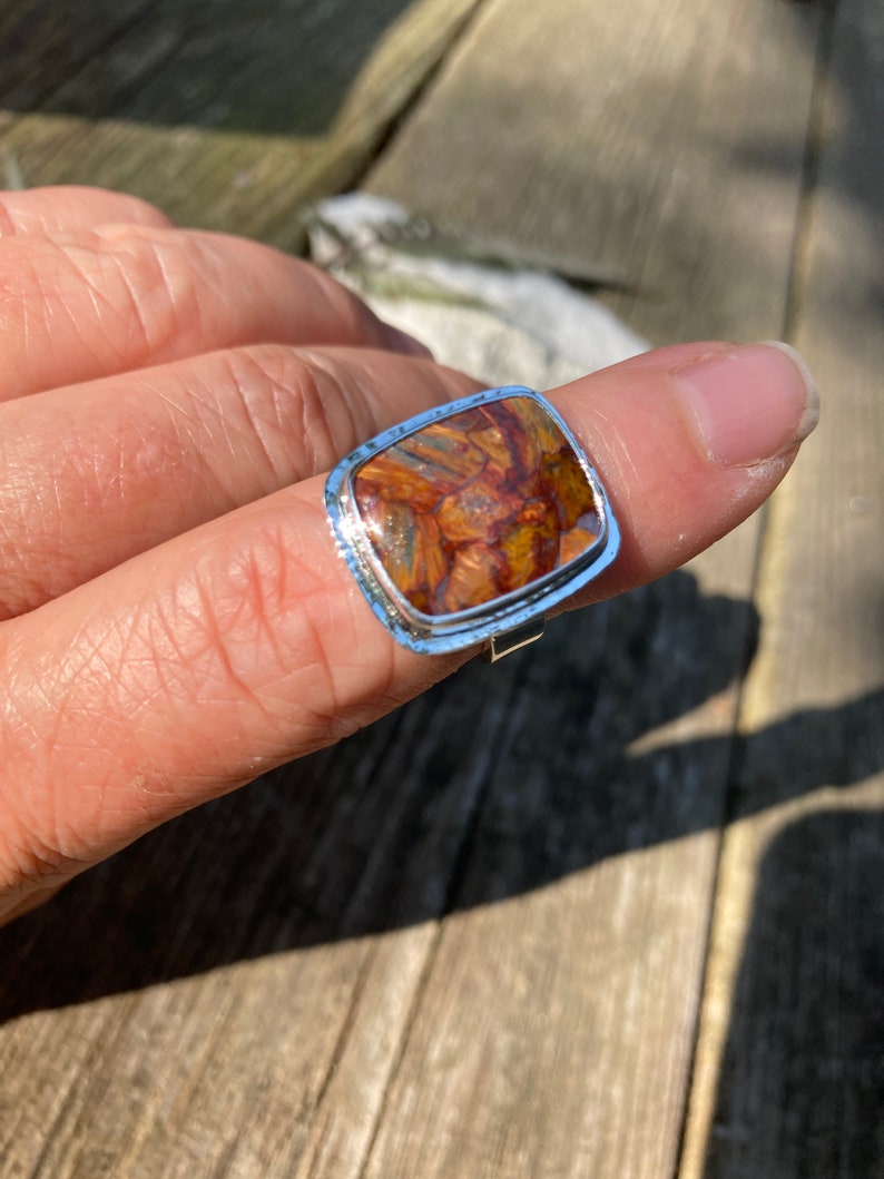 May include: A silver ring featuring a square-shaped, polished gemstone with a marbled pattern of deep brown, amber, and golden-orange hues. The metal setting has a textured, hammered edge surrounding the stone, creating a distinct border.