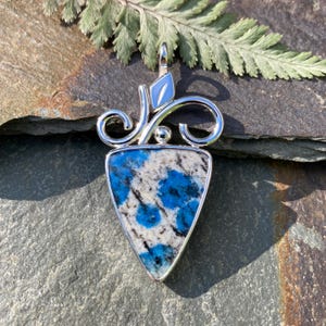 May include: A triangular pendant featuring a polished stone with vibrant blue and white speckled patterns. The stone is set in a silver-toned metal frame with decorative swirling wire accents and a small leaf-shaped detail at the top bail.