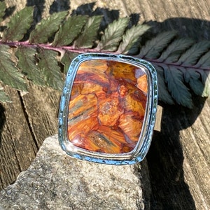 May include: A rectangular ring featuring a polished, multi-coloured stone with swirling patterns of orange, brown, and grey. The stone is set in a silver-toned metal frame with a textured, hammered edge, creating a distinct border around the centre.