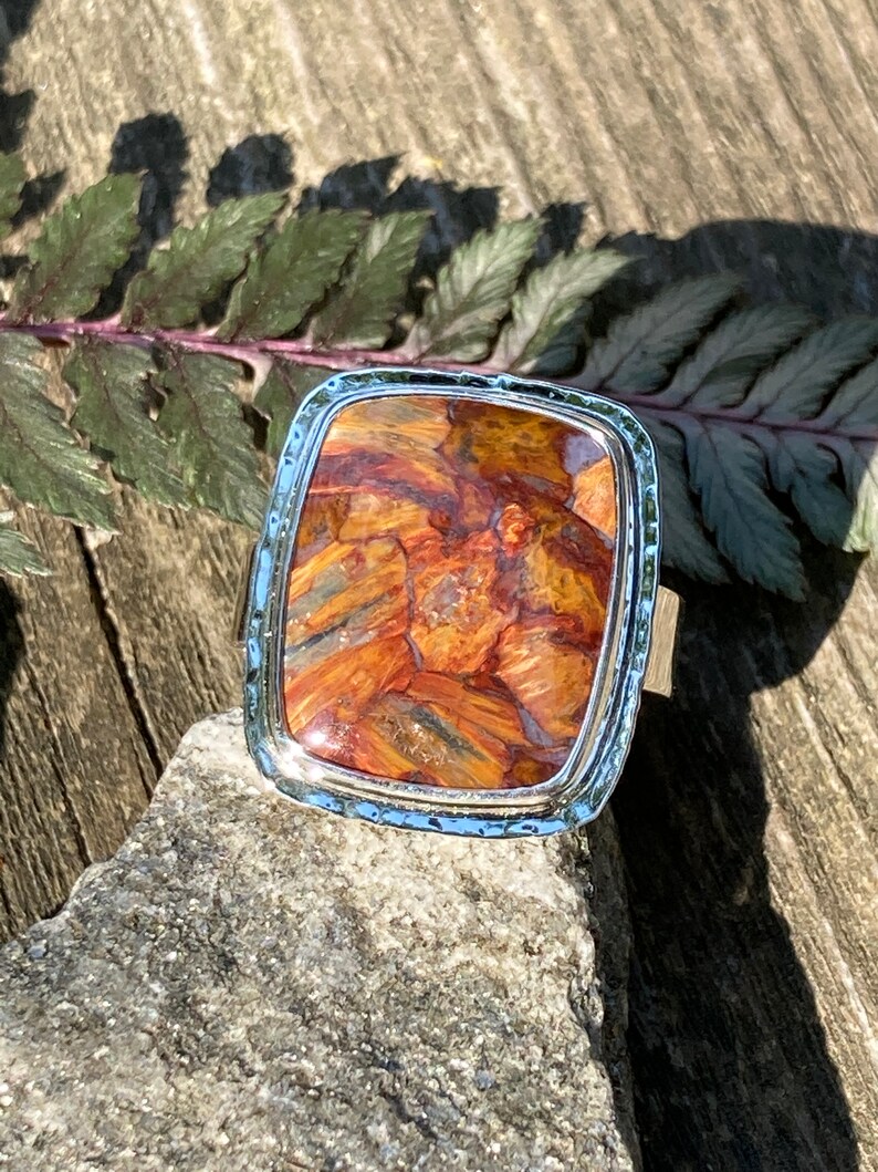 May include: A rectangular ring featuring a polished, multi-coloured stone with swirling patterns of orange, brown, and grey. The stone is set in a silver-toned metal frame with a textured, hammered edge, creating a distinct border around the centre.