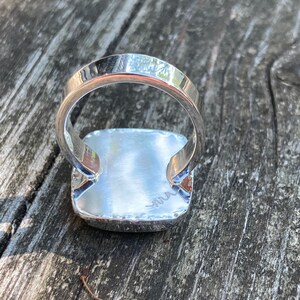 May include: A silver ring featuring a square-shaped setting with a polished, reflective surface. The band is smooth and rounded, and the underside of the square setting displays a small, engraved signature mark consisting of a stylised, wavy line.