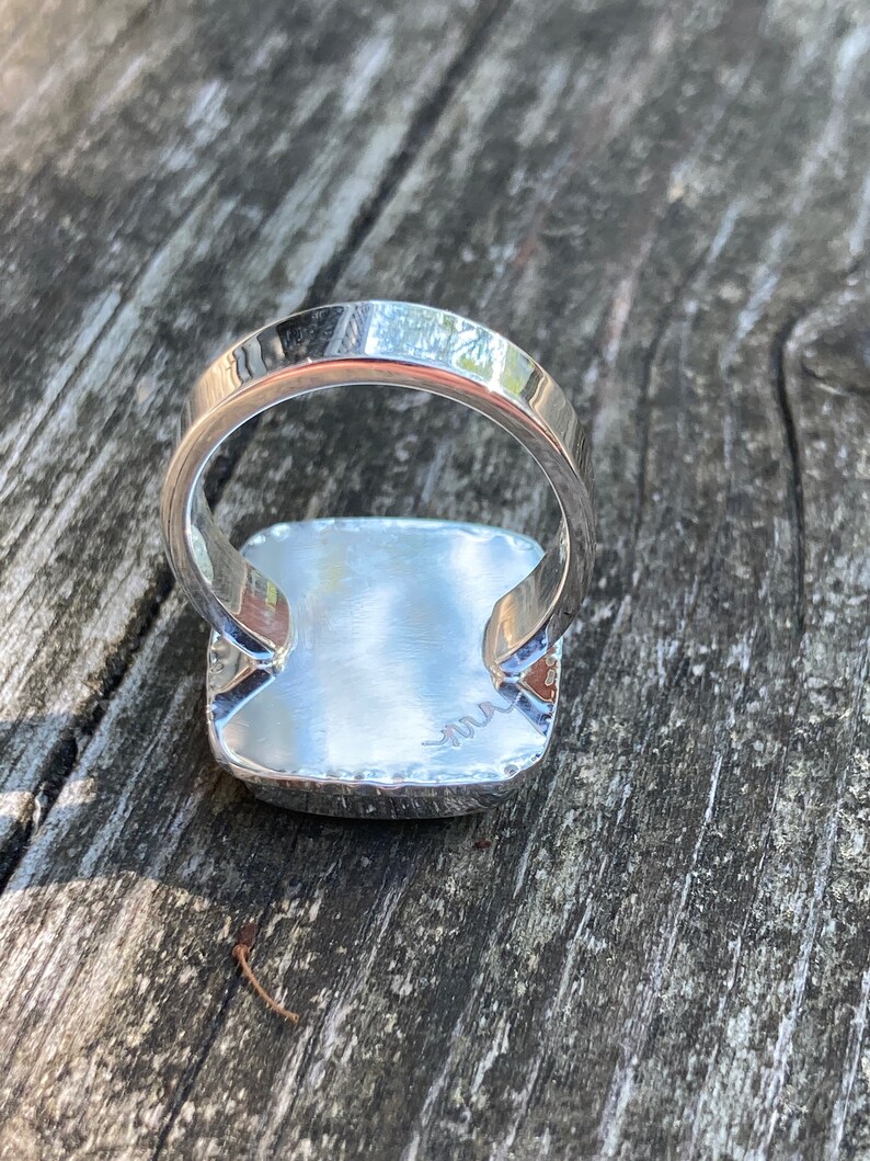 May include: A silver ring featuring a square-shaped setting with a polished, reflective surface. The band is smooth and rounded, and the underside of the square setting displays a small, engraved signature mark consisting of a stylised, wavy line.