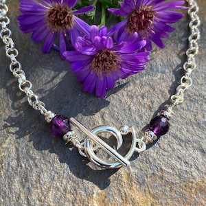 May include: A silver chain necklace featuring a toggle clasp closure. The necklace is accented with two round, faceted purple amethyst beads positioned on either side of the central silver toggle ring and bar mechanism, set against purple flowers.
