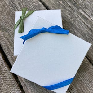 May include: Two square white gift boxes with textured surfaces. The top box is wrapped with a bright blue raffia ribbon tied in a bow. The bottom box is partially visible and features a green raffia ribbon tied in a bow on its top corner.