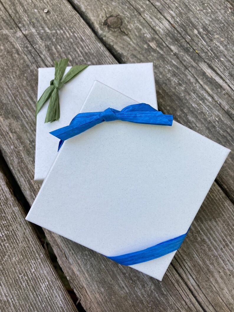 May include: Two square white gift boxes with textured surfaces. The top box is wrapped with a bright blue raffia ribbon tied in a bow. The bottom box is partially visible and features a green raffia ribbon tied in a bow on its top corner.