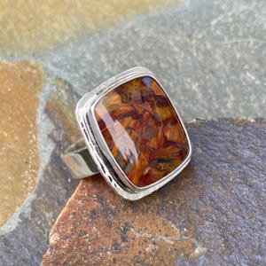 May include: A silver ring featuring a rectangular, polished gemstone with a vibrant, multi-tonal pattern of deep orange, brown, and golden yellow streaks. The metal setting has a smooth, reflective finish and a simple, sturdy band design.