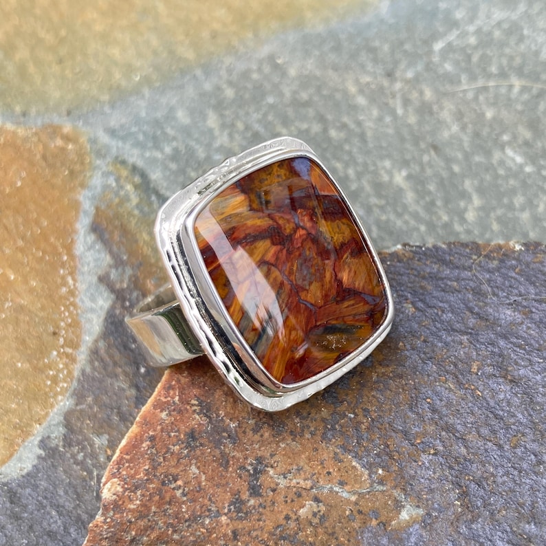 May include: A silver ring featuring a rectangular, polished gemstone with a vibrant, multi-tonal pattern of deep orange, brown, and golden yellow streaks. The metal setting has a smooth, reflective finish and a simple, sturdy band design.