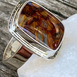 May include: A silver ring featuring a rectangular, polished gemstone with a marbled pattern of deep brown, golden yellow, and black tones. The stone is set in a smooth, reflective silver bezel with a wide, solid silver band that tapers slightly.