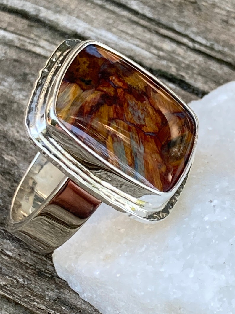 May include: A silver ring featuring a rectangular, polished gemstone with a marbled pattern of deep brown, golden yellow, and black tones. The stone is set in a smooth, reflective silver bezel with a wide, solid silver band that tapers slightly.