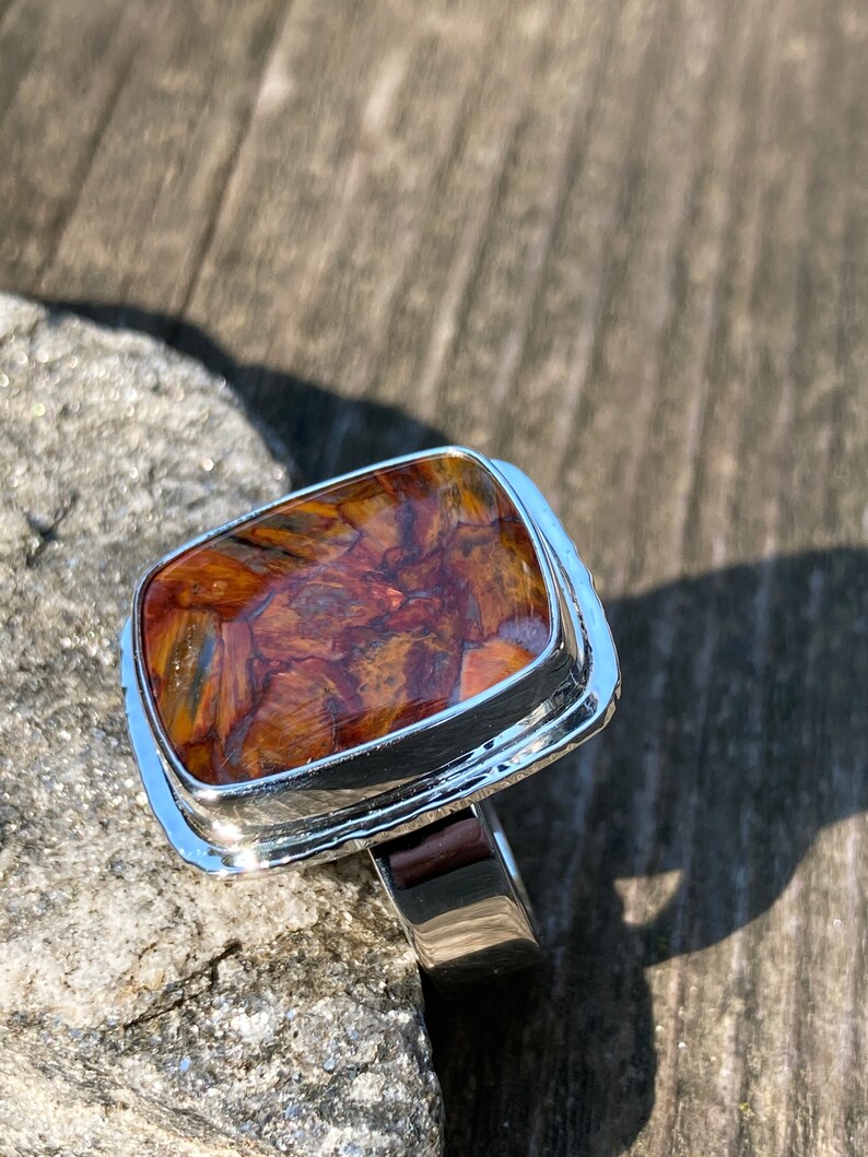 May include: A silver ring featuring a rectangular, polished pietersite gemstone with swirling patterns of deep brown, gold, and blue. The stone is set in a smooth, reflective silver bezel with a wide, solid silver band that tapers slightly.