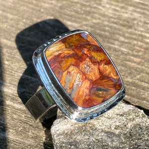May include: A silver ring featuring a rectangular, polished gemstone with a complex pattern of deep amber, rust-red, and golden-brown streaks. The metal setting has a smooth, reflective finish with a slightly hammered texture along the outer edge.
