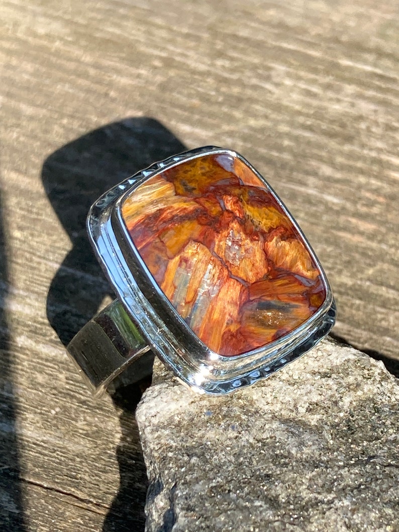 May include: A silver ring featuring a rectangular, polished gemstone with a complex pattern of deep amber, rust-red, and golden-brown streaks. The metal setting has a smooth, reflective finish with a slightly hammered texture along the outer edge.