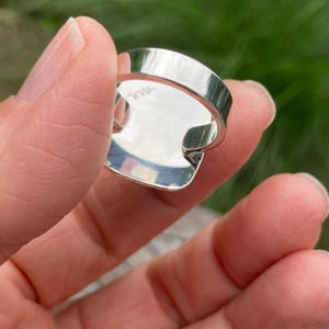 May include: A polished silver ring featuring a smooth, reflective band and a flat, circular setting. The interior of the band displays a small, engraved signature or mark, showcasing the metallic lustre and clean construction of the jewellery piece.