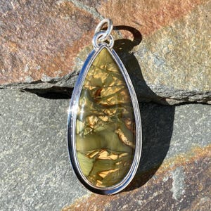 May include: A teardrop-shaped pendant featuring a polished, multi-coloured stone with swirling patterns of olive green, tan, and dark brown, encased in a smooth, reflective silver-toned metal bezel setting with a circular bail at the top.