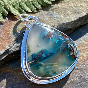May include: A teardrop-shaped pendant featuring a polished gemstone with translucent white, deep teal, and rust-coloured inclusions. The stone is encased in a textured sterling silver bezel setting with a matching silver jump ring at the top.